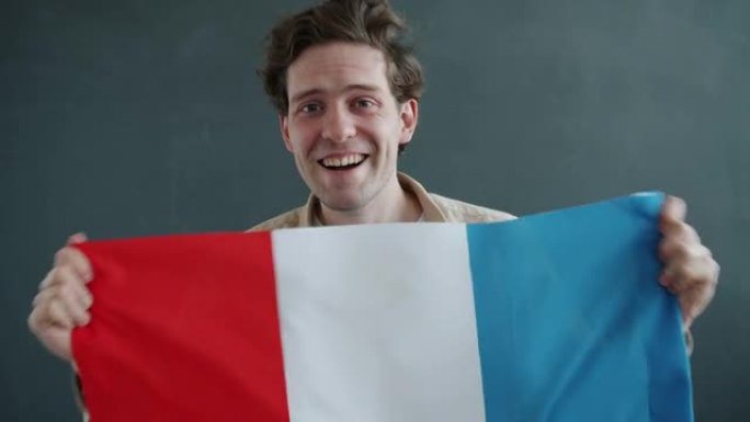快乐的年轻人拿着法国国旗微笑着看着灰色背景上的相机的慢动作肖像