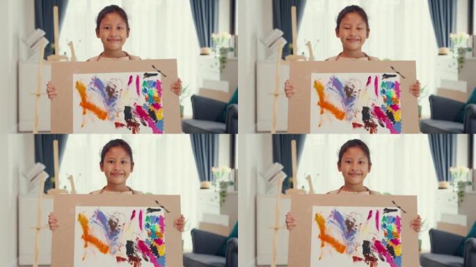 亚洲蹒跚学步的女孩，围裙站在前面的沙发椅上举起她的艺术作品自然甜美的微笑看着相机在家里的客厅展示抽象