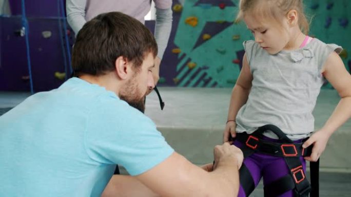 友好的攀岩教练在健身房室内帮助儿童使用安全带