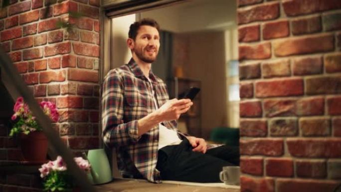 快乐英俊的白人男子坐在舒适公寓的窗台上使用智能手机。穿着休闲衬衫的家伙浏览互联网，在社交媒体上查看视