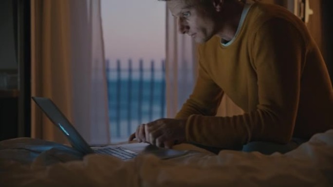 晚上，棕色短发的男人在笔记本电脑上工作，坐在酒店房间的床上，背景是阳台和大海，使用智能手机