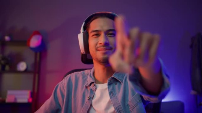 快乐亚洲男玩家戴着耳机参与玩视频游戏彩色霓虹灯电脑在客厅晚上现代房子。