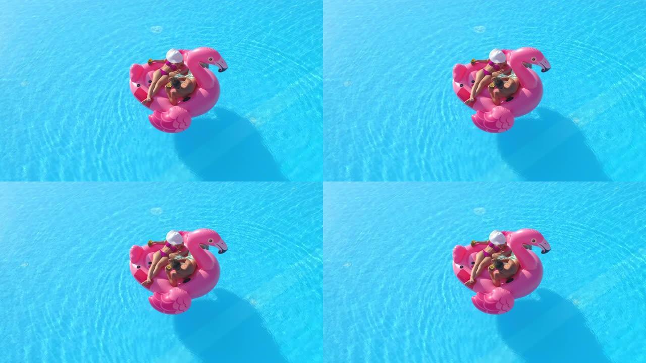 空中: 年轻的旅游夫妇在泳池派对上躺在粉红色的火烈鸟漂浮物上