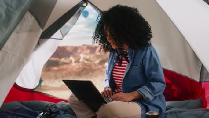 在落基峡谷顶部的帐篷里，用笔记本电脑在非洲发型的年轻多民族女性旅行者兴奋不已。冒险的山地徒步旅行者生