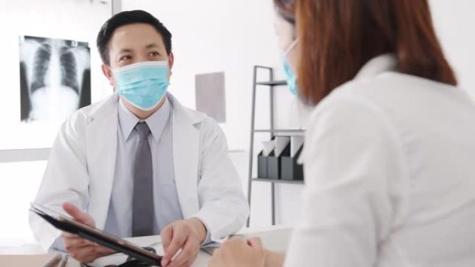 严重的亚洲男性医生戴防护口罩使用平板电脑正在传递重大新闻谈话与医院办公室的女性患者讨论结果或症状。