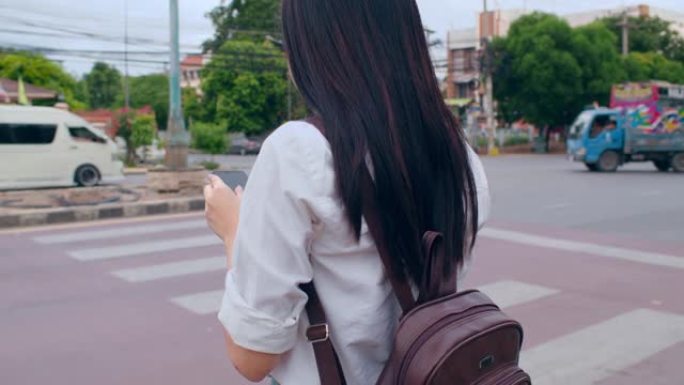 年轻的亚洲背包客女游客用相机站在十字路口街前看手机搜索导航应用的位置。