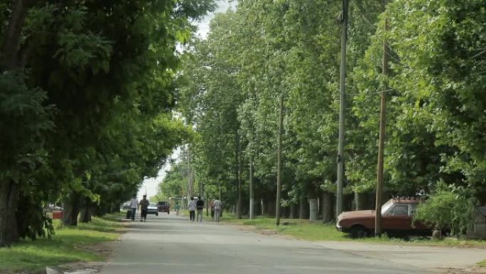 在阿根廷，乡间小路有树木通向远处，一辆旧车停在一边，人们在散步。