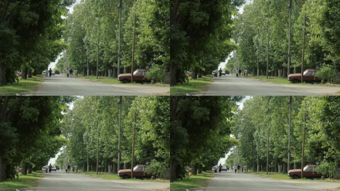 在阿根廷，乡间小路有树木通向远处，一辆旧车停在一边，人们在散步。