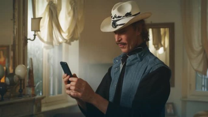 戴着牛仔帽的无忧无虑的中年快乐男人的电影镜头正在使用智能手机，在家中客厅的相机中微笑。