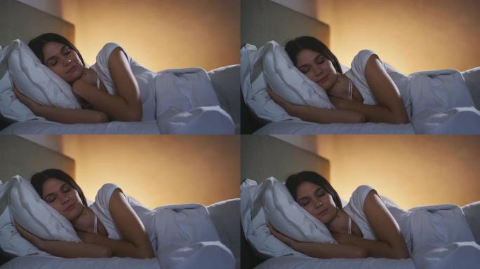 一名年轻的黑发女子在舒适的床上安详地睡在温暖的羽绒被毯子下，卧室里开着夜灯。舒适、放松、睡眠、健康、