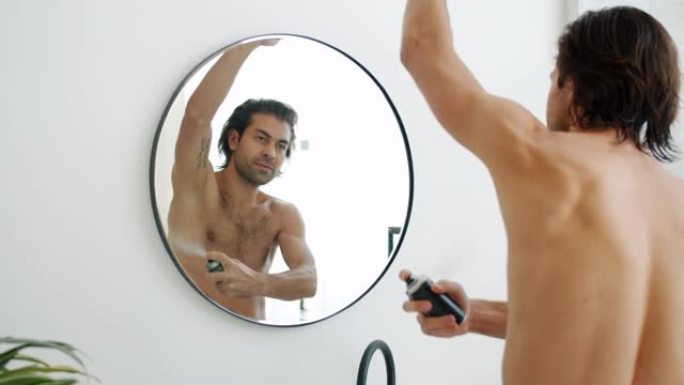 英俊的年轻人在浴室使用除臭喷雾在腋窝上照镜子