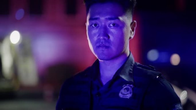 特写:一名年轻的亚洲男警察在警笛灯光下摆姿势，坚定地看着镜头。英勇的执勤警官，保护平民，维护正义，打