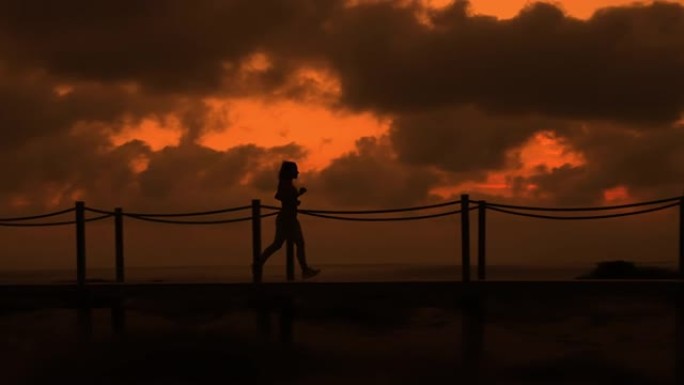 慢动作，在深橙色的日落中，无法辨认的女性在海滩上的木板路上奔跑的照片