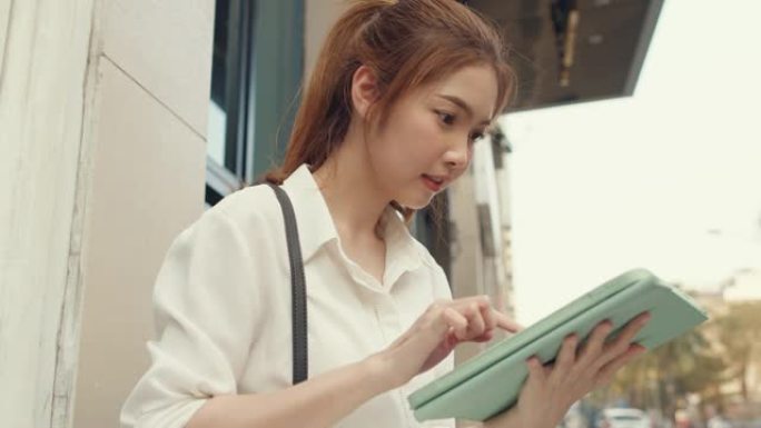 在城市现代城市中独自一人坐在户外时，穿着时尚办公服装的成功年轻亚洲女商人使用数字平板电脑和打字短信。