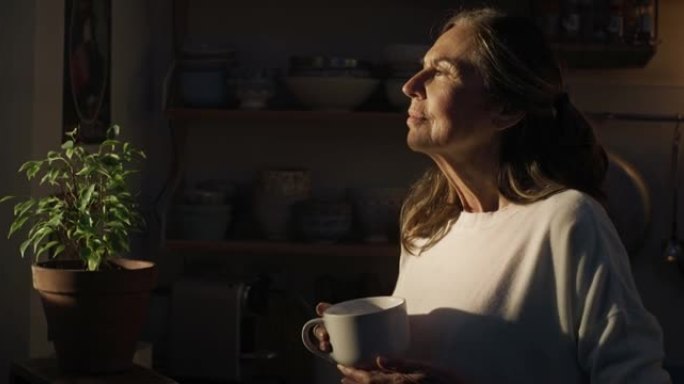 快乐微笑的高级养老金领取者的电影镜头正在喝热茶或美式咖啡，早晨在家里的厨房里透过阳光透过窗户看。舒适