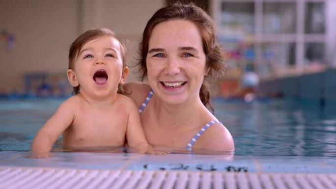 妈妈抱着可爱的婴儿在室内游泳池里看着微笑