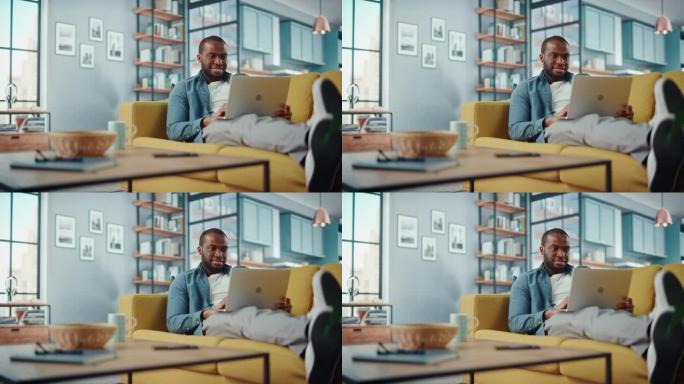 英俊的黑人非洲裔美国男子躺在舒适的客厅的沙发上，在笔记本电脑上工作。自由职业者在家工作。浏览互联网，
