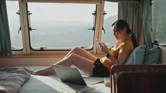 年轻的亚洲女性使用智能手机在房车露营期间远程工作前在卧室放松。
