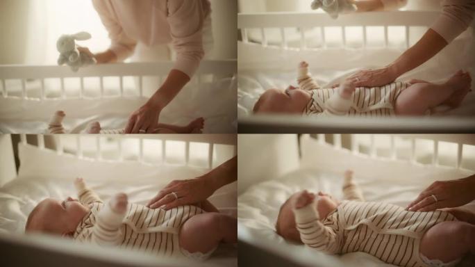 快乐的母亲在舒适温暖的房间里和一个躺在婴儿床背上的可爱的新生婴儿玩耍。妈妈拿着一个玩具在一个幼儿的旁