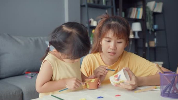 快乐快乐的亚洲家庭妈妈教蹒跚学步的女孩油漆陶瓷锅在客厅的桌子上放松。