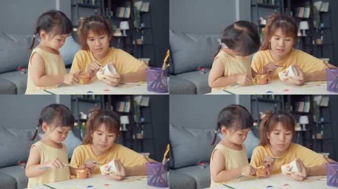 快乐快乐的亚洲家庭妈妈教蹒跚学步的女孩油漆陶瓷锅在客厅的桌子上放松。