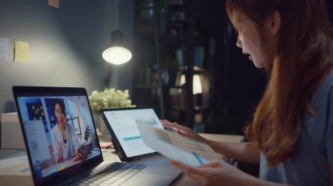 使用笔记本电脑的亚洲女性与同事谈论视频通话中的工作，而晚上在家在客厅工作。自我隔离，社会疏远，冠状病