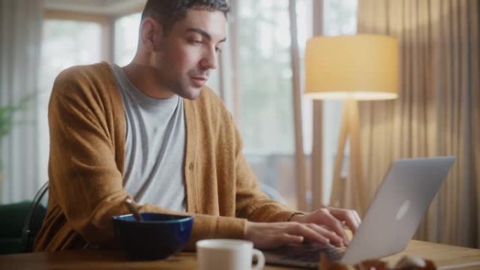 英俊的成年男子使用笔记本电脑，坐在厨房里，在公寓里吃早餐粥或麦片。有魅力的男人正在网上购物，在流媒体