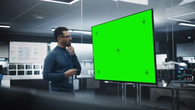 多种族的首席软件工程师独自站在会议室中，使用带有绿屏模拟模板的大型电视显示屏，其中包含开发部门创建的