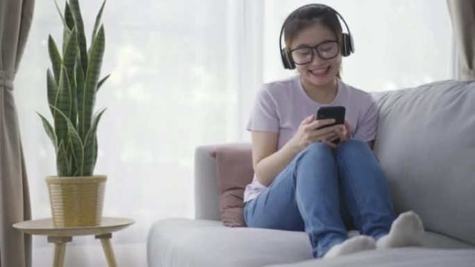 年轻的亚洲女性在沙发上放松，在家里的客厅用耳机听音乐