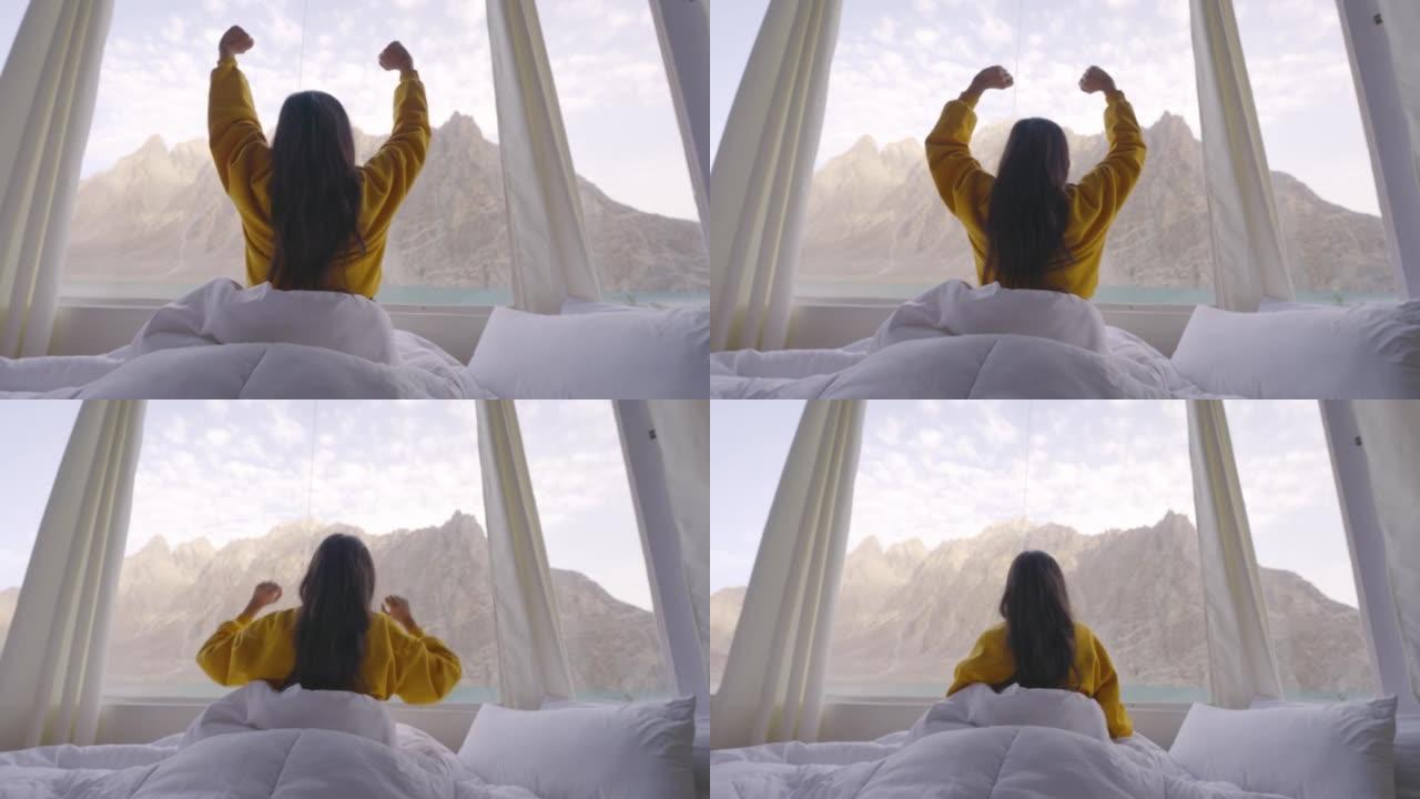 穿着黄色衣服，裹着温暖的毯子的年轻女子的后视图在早晨醒来，在床上透过湖边的窗户伸张。种族多样化的旅行