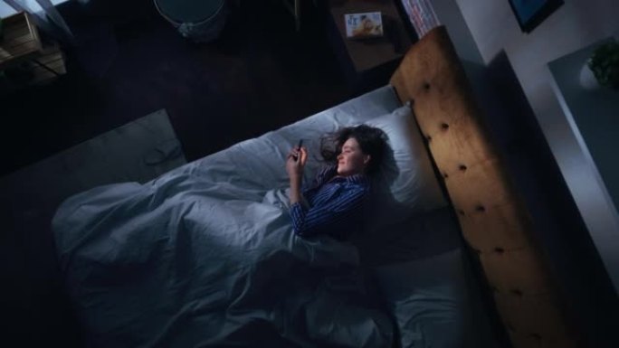 顶景卧室公寓: 美丽的年轻女子晚上在床上使用智能手机。微笑的女孩浏览社交媒体，约会应用中的浪漫，远程