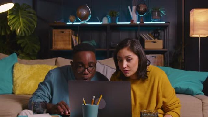 两个快乐的年轻多民族学生朋友学习，使用坐在客厅地板上的笔记本电脑一起在线工作。