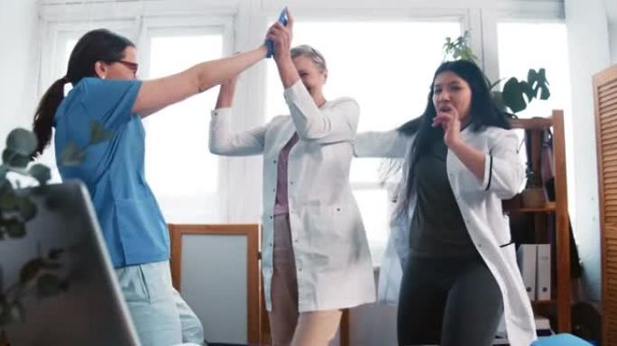 战胜冠状病毒。三名快乐兴奋的多民族女医生在诊所办公室庆祝巨大的成功跳舞