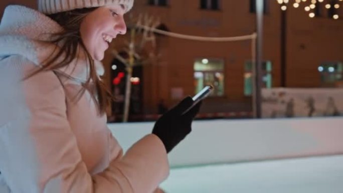快乐的年轻女子晚上在溜冰场上滑冰时使用手机