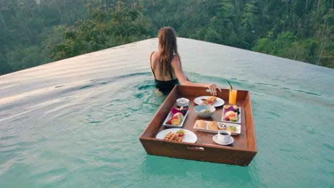 旅行女人在游泳池吃早餐，在豪华酒店水疗中心享受异国美食，日出时可以看到热带丛林