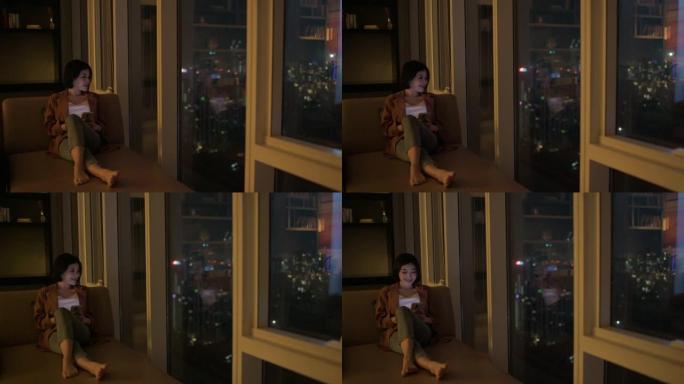 亚洲中国美女晚上在沙发上的客厅用手机靠近城市的窗户
