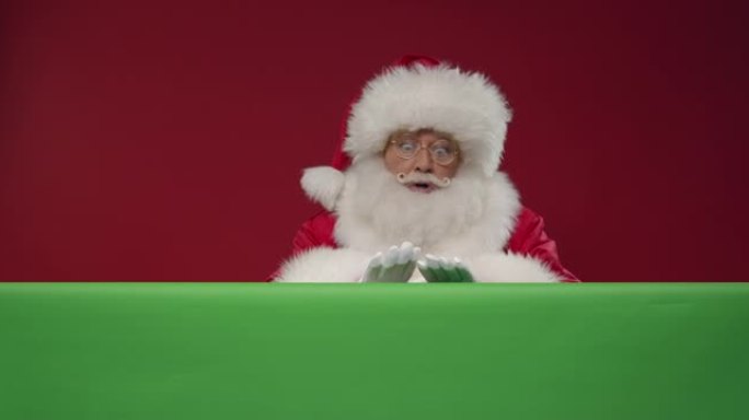 快乐的圣诞老人从绿色屏幕后面出现，就像一个小丑从红色背景上的盒子里出来，显示拇指向上的标志，然后隐藏