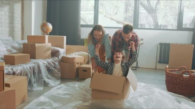快乐的父母有乐趣推动兴奋可爱的男孩在纸板箱在地板上庆祝在新的宽敞的家。