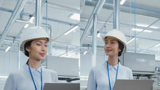 在电子制造工厂，一名戴着安全帽行走和使用笔记本电脑的亚洲女工程师的垂直肖像。处理日常任务和研发数据的