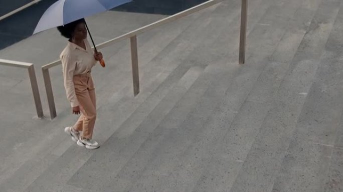 一位年轻迷人的黑皮肤女商人穿着米色衣服，打着雨伞走上楼梯