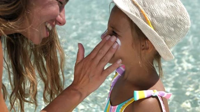 一个小女孩的肖像在海滩上笑着，而她的母亲在她的脸上涂了防晒霜。暑假期间，年轻女子保护女儿的皮肤免受阳