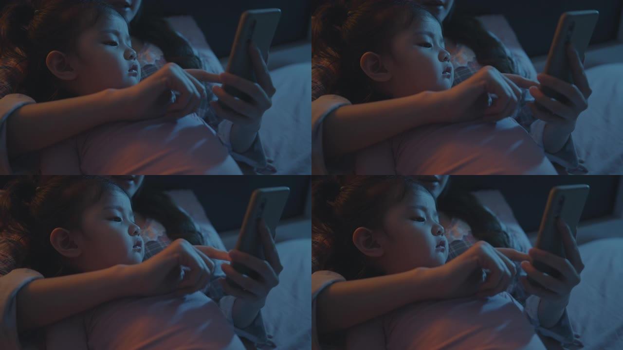 迷人可爱的儿童女孩晚上和妈妈在床上看智能手机。