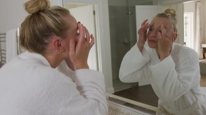 穿着浴袍的白人妇女在浴室照镜子时抚摸着脸