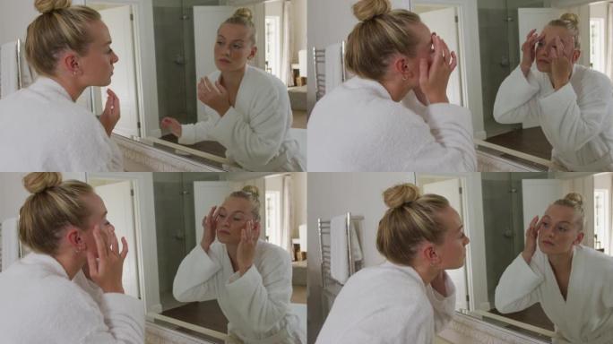 穿着浴袍的白人妇女在浴室照镜子时抚摸着脸
