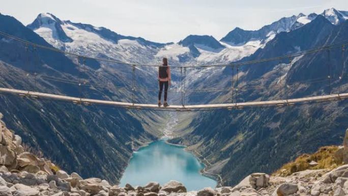 女徒步旅行者站在山涧上的吊桥上，欣赏山谷湖和周围山脉的壮丽景色