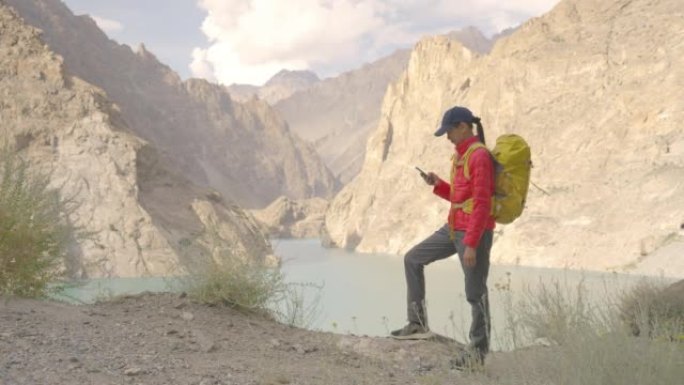自由快乐的旅行者年轻女性穿着红色的冬季夹克，背着黄色背包站着使用手机享受湖边的旅行假期。种族多样化的
