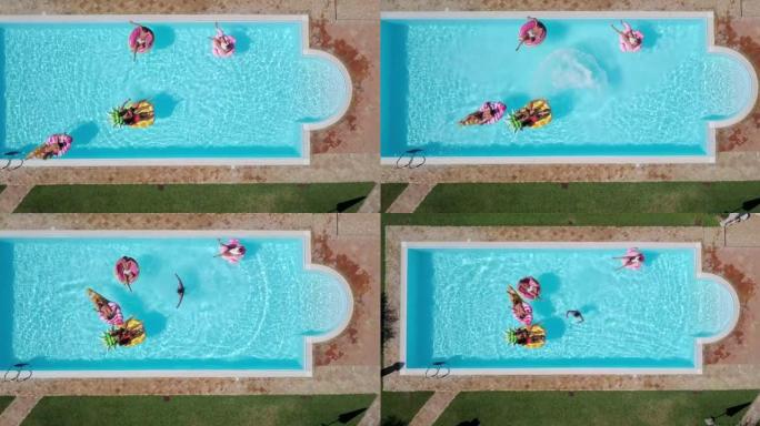 在阳光明媚的日子里，穿着泳衣的年轻多民族朋友的空中拍摄正在游泳池里放松，那里有五颜六色的充气设备。