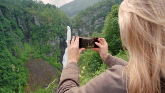 4k视频片段，一个无法识别的女人站着并用手机拍摄挪威瀑布的照片