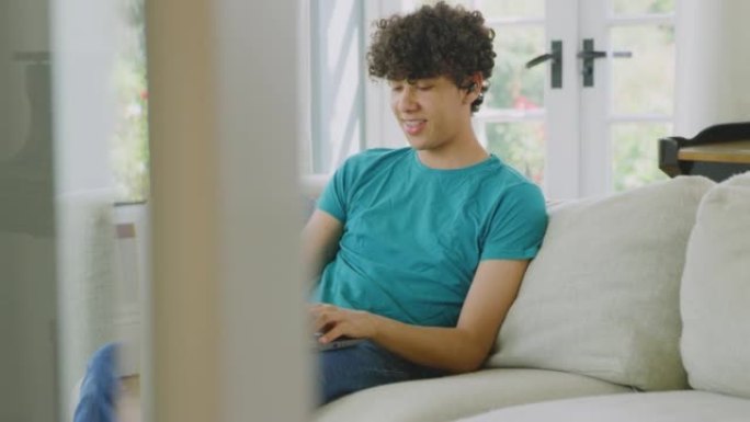 十几岁的男孩戴着无线耳塞坐在家里的沙发上听音乐或在笔记本电脑上播放电影-慢动作拍摄