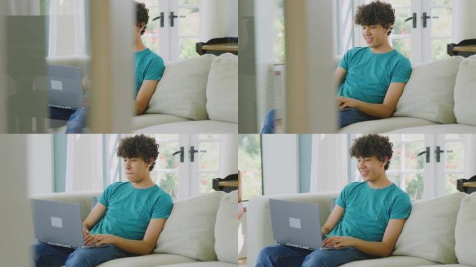 十几岁的男孩戴着无线耳塞坐在家里的沙发上听音乐或在笔记本电脑上播放电影-慢动作拍摄